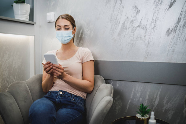 Una joven atractiva que usa mascarilla facial y su teléfono celular mientras está sentada en una silla en la sala de espera en la oficina del dentista. - Foto, imagen