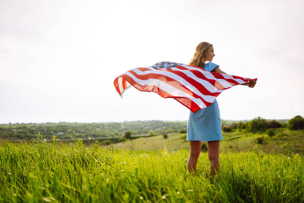 Young woman with american flag on blooming meadow. 4th of July. Independence Day. Patriotic holiday. USA flag fluttering in the wind. - Foto, Imagen