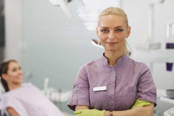 Portret van een lachende vrouwelijke tandarts die met gevouwen armen naar de camera in haar kantoor kijkt. - Foto, afbeelding