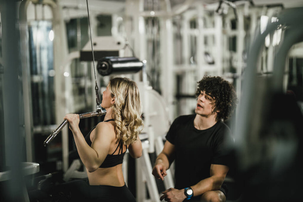 Eine junge muskulöse Frau trainiert im Fitnessstudio an einer Maschine, die von einem Trainer unterstützt wird. - Foto, Bild