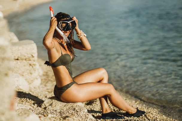 Viehättävä nuori nainen istuu veden äärellä sukellusnaamionsa ja snorkelinsa kanssa ja valmistautuu snorklaamaan meressä.. - Valokuva, kuva