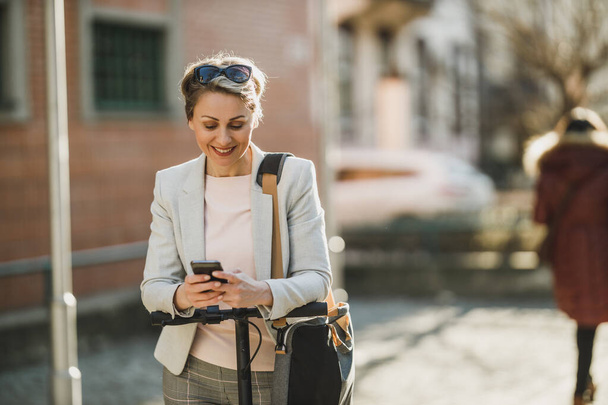 Dojrzała bizneswoman korzystająca ze smartfona podczas podróży skuterem elektrycznym przez miasto. - Zdjęcie, obraz