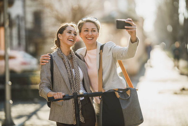 Zwei erfolgreiche Geschäftsfrauen machen ein Selfie mit dem Smartphone, während sie durch die Stadt laufen. - Foto, Bild