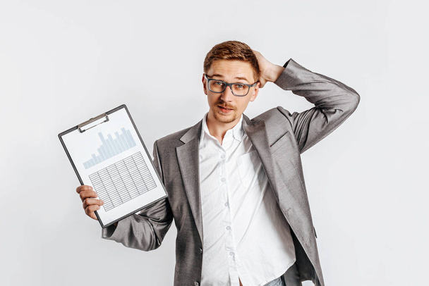 Young handsome brunet man in glasses in a suit is unhappy with business documents and charts on a white isolated background - Foto, afbeelding