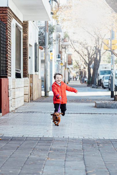 Happy little boy running with his dachshund dog on a city sidewalk. - Photo, image