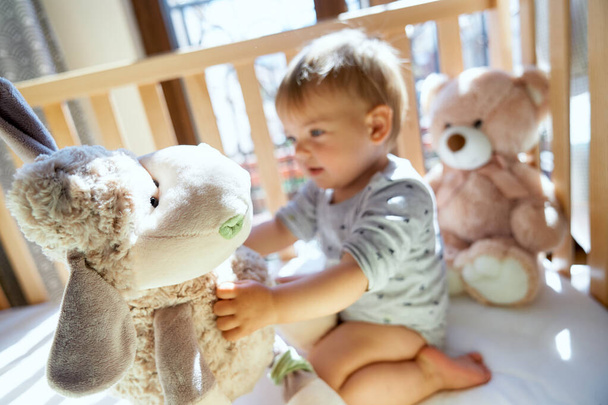 Улыбающийся маленький ребенок сидит на деревянной кровати и держит большую плюшевую овцу - Фото, изображение