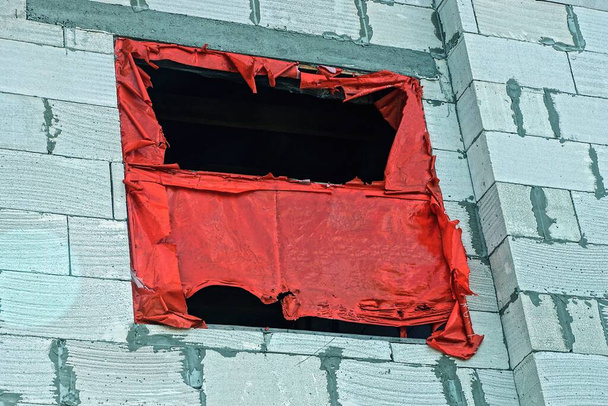 ένα παράθυρο κλειστό με σχισμένο κόκκινο σελοφάν σε έναν λευκό τοίχο από τούβλα ενός μισοτελειωμένου σπιτιού στο δρόμο - Φωτογραφία, εικόνα