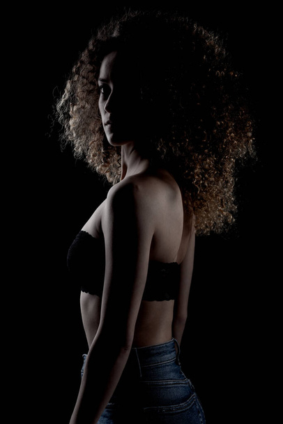 Side lit brunette girl with long curly hair, silhouette studio portrait on dark background. - Foto, afbeelding