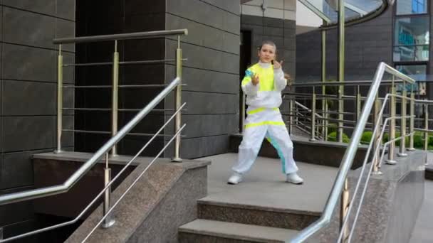 Een meisje in een stijlvol pak danst een HIP-HOP dans bij de ingang van het kantoorgebouw bij de metalen leuningen. Moderne dans in een stedelijke straatomgeving. - Video