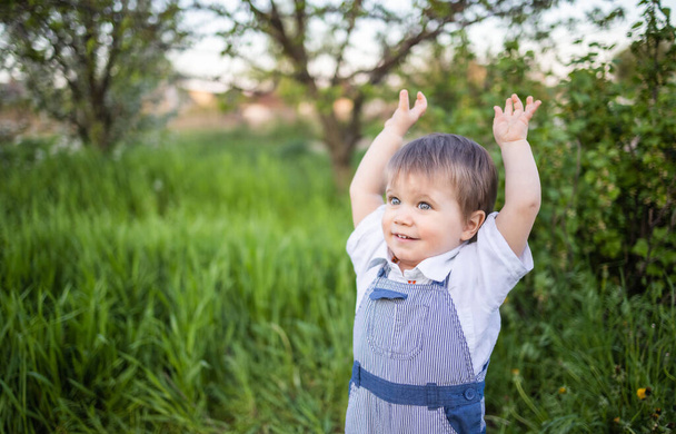 Cute boy in fashionable clothes with blue eyes plays and jumps in the tall grass in a large green blooming garden - Foto, imagen