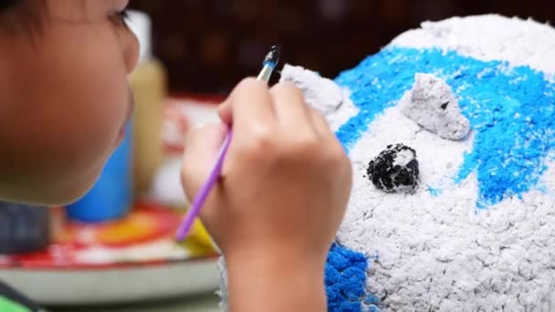 Маленька дівчинка малює пензлем на фігурі білої свинячої пап'є-маше вдома. Зробіть свій власний скарбничку
. - Кадри, відео