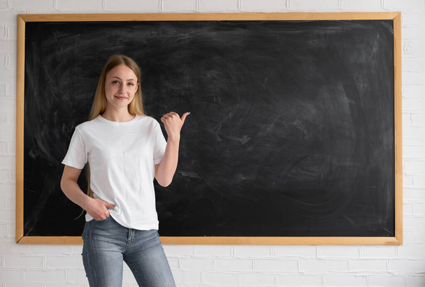 Egy fiatal lány diák vagy tanár áll a kréta tábla mellett mosolyogva. A hüvelykujjával üres helyet jelez a szövegnek.. - Fotó, kép