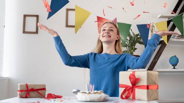 A blonde Caucasian girl sitting at a festive table on her birthday laughs and throws up different confetti with her hands. Woman in a festive hat at a party at home. Remote birthday concept. - Photo, Image