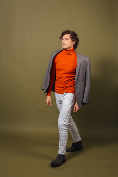 Full length portrait of a tall handsome man dressed in orange turtleneck, grey jacket and white jeans posing on the green background - Valokuva, kuva