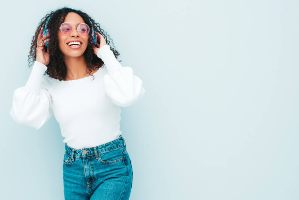 Beautiful black woman with afro curls hairstyle.Smiling model in sweater and jeans.Sexy carefree female listening music in wireless headphones.Posing in studio near light blue wall - Φωτογραφία, εικόνα