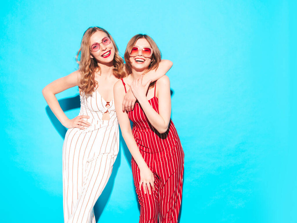 Deux jeunes belles hipsters souriantes en salopette d'été tendance. Sexy femmes insouciantes posant près du mur bleu en studio. Modèles à la mode et positifs s'amuser. Joyeux et heureux - Photo, image