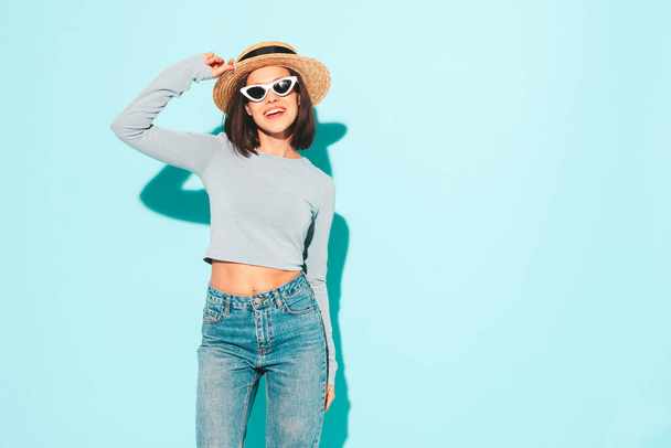 Portrait of young beautiful smiling female in trendy summer hipster  clothes. Sexy carefree woman posing near blue wall in studio. Positive model having fun in hat. Cheerful and happy in sunglasses - Fotó, kép