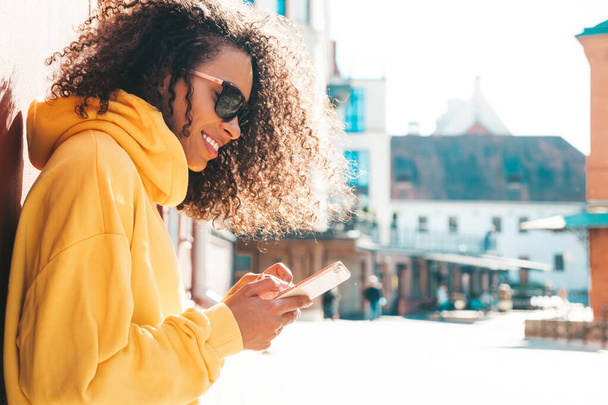 Beautiful black woman with afro curls hairstyle.Smiling model in yellow hoodie. Sexy carefree female posing on the street background in sunglasses. Looking at smartphone screen, using apps - Photo, image