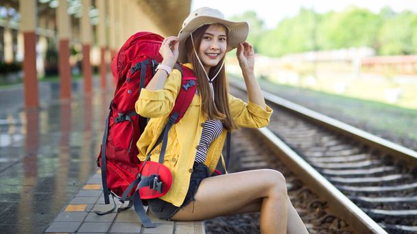 Seitenansicht einer Backpackerin, die lächelt und vor die Kamera klebt, während sie am Bahnhof sitzt  - Foto, Bild