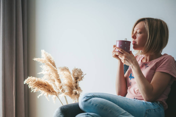 Giovane donna che beve bevande calde su un divano a casa. Sta annusando un caffe 'dalla tazza.. - Foto, immagini
