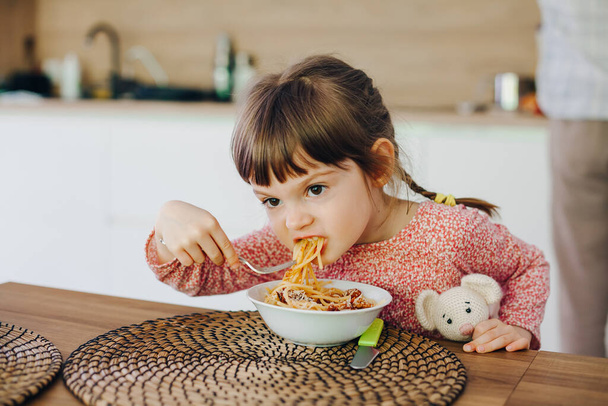 Cute little girl eating spaghetti with sauce sitting in a kitchen at home - Photo, Image