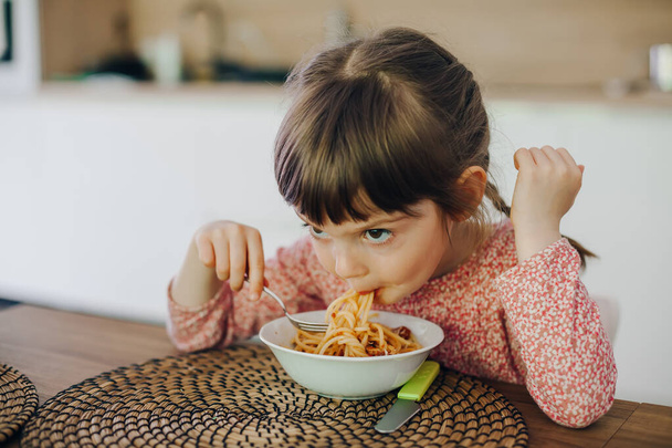 Cute little girl eating spaghetti with sauce sitting in a kitchen at home - Φωτογραφία, εικόνα