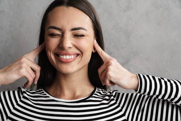 Close up of a smiling young attractive woman blocking her ears isolated over gray background - Foto, Imagen