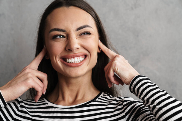 Close up of a smiling young attractive woman blocking her ears isolated over gray background - Foto, imagen