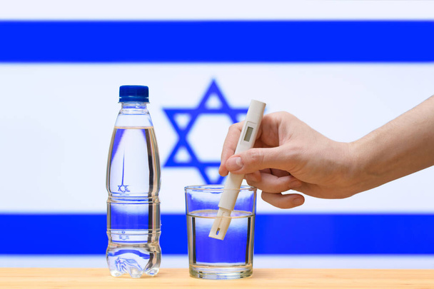 Una mano con un tester dell'acqua effettua una misurazione in un bicchiere di acqua limpida sullo sfondo della bandiera di Israele. Test e valutazione dell'approvvigionamento di acqua potabile in Israele. - Foto, immagini