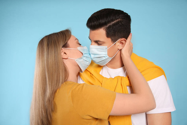 Couple in medical masks trying to kiss on light blue background - Fotoğraf, Görsel