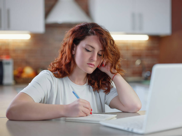 Egy gyönyörű, fiatal, vörös hajú nő ül az asztalnál a konyhában a laptopjával és jegyzetel. tanulói távoktatás, házi feladat, videólecke hallgatása. Távoktatási koncepció - Fotó, kép