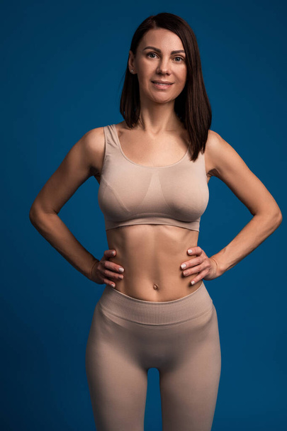 Woman in sports top and leggings with arms akimbo on blue background - Photo, Image