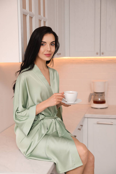 Pretty young woman wearing beautiful silk robe with cup of coffee in kitchen - Foto, immagini