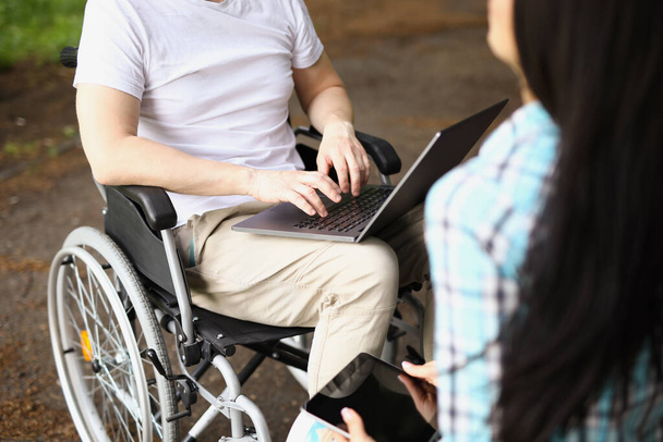 Мужчина в инвалидной коляске работает на ноутбуке на улице рядом с женщиной сидит и держит планшет - Фото, изображение