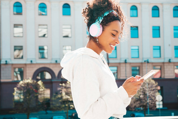 Beautiful black woman with afro curls hairstyle.Smiling model in yellow hoodie.Sexy carefree female enjoying listening music in wireless headphones.Posing on street background at sunset.Holds phone - Photo, Image