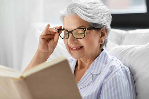 oude vrouw in bril lezen boek in bed thuis - Foto, afbeelding