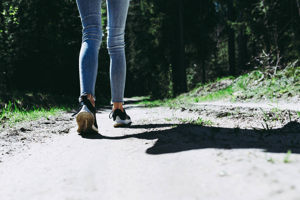 Widok z tyłu zdjęcie kobiecych nóg turystycznych z trampkami spacerującymi po leśnej drodze.Summer day sunshine. - Zdjęcie, obraz