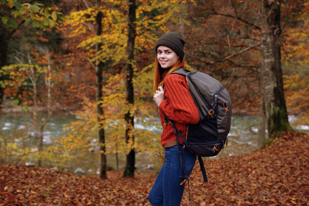 woman in autumn park with fallen leaves and backpack on her back river in the background - Photo, Image