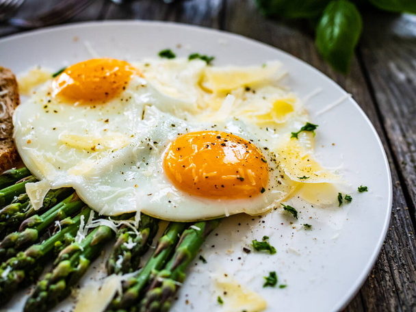 Tasty breakfast. Sunny side up eggs with green asparagus and parmesan served on black plate on wooden table - Photo, image