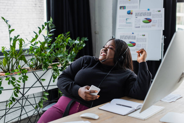 boldog afro-amerikai plusz méret operátor headset mikrofon beszél és gazdaság okostelefon az irodában  - Fotó, kép