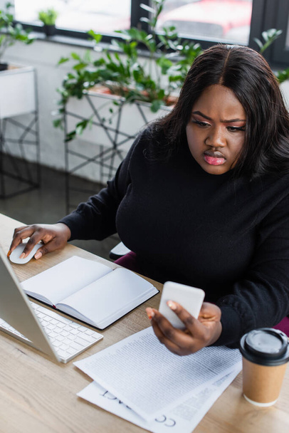 africano americano más tamaño mujer de negocios mirando teléfono inteligente cerca de monitor de computadora en la oficina  - Foto, Imagen