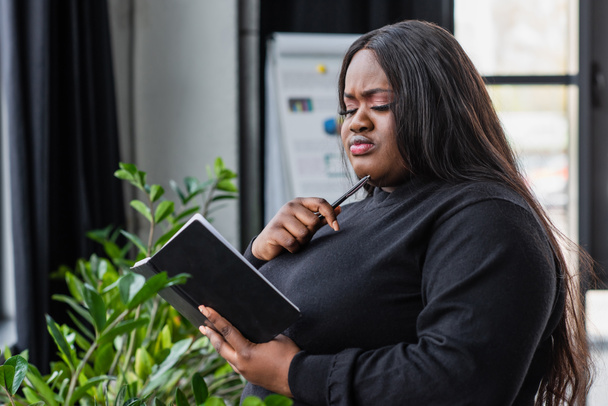 pensive african american plus size businesswoman holding notebook in office - Foto, Bild
