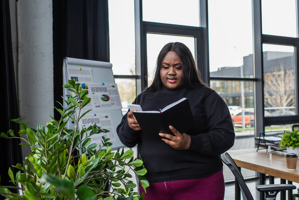 african american plus size businesswoman holding notebook and smartphone in office - Fotoğraf, Görsel