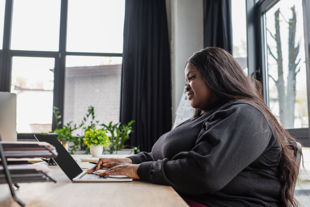 happy african american plus size woman typing on laptop keyboard in office - Fotó, kép