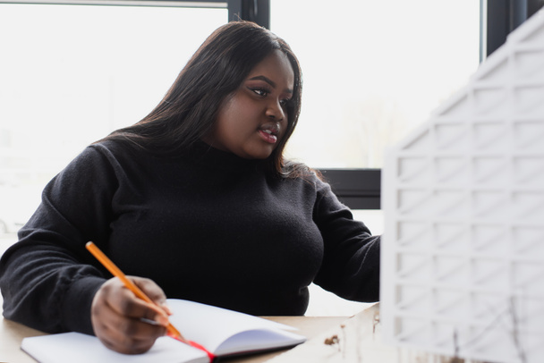 afroamericano más tamaño diseñador escritura en cuaderno mientras mira modelo de casa  - Foto, Imagen