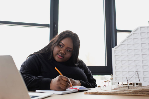Afrikanische amerikanische Plus-Size-Designerin schreibt in Notizbuch, während sie Laptop in der Nähe des Hausmodells betrachtet  - Foto, Bild