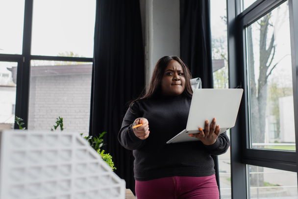 Afrikaans amerikaans plus size vrouw houden laptop en wijzen met potlood op huis model  - Foto, afbeelding