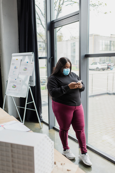 afro-amerikai plusz méret üzletasszony orvosi maszk segítségével okostelefon közelében elmosódott ház modell  - Fotó, kép