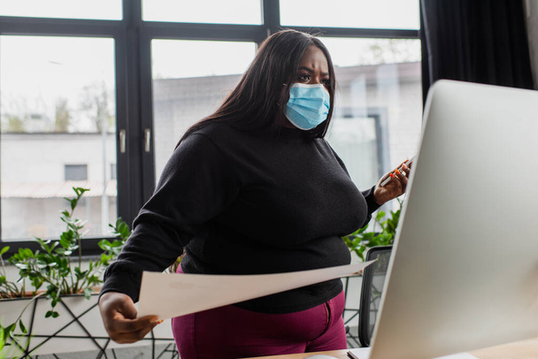 Afrykańska amerykanka plus size businesswoman w masce medycznej trzymając smartfon i papier patrząc na monitor  - Zdjęcie, obraz