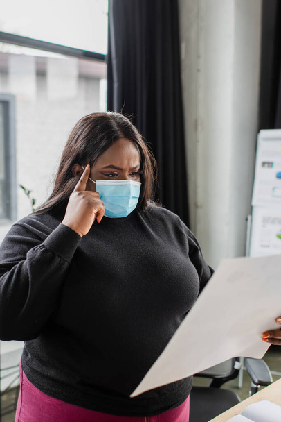 african american plus size businesswoman in medical mask holding paper in office - Photo, Image
